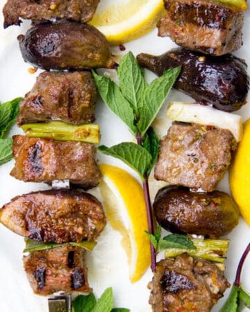 Two grilled lamb and fig skewers surrounded by lemon slices and mint sprigs