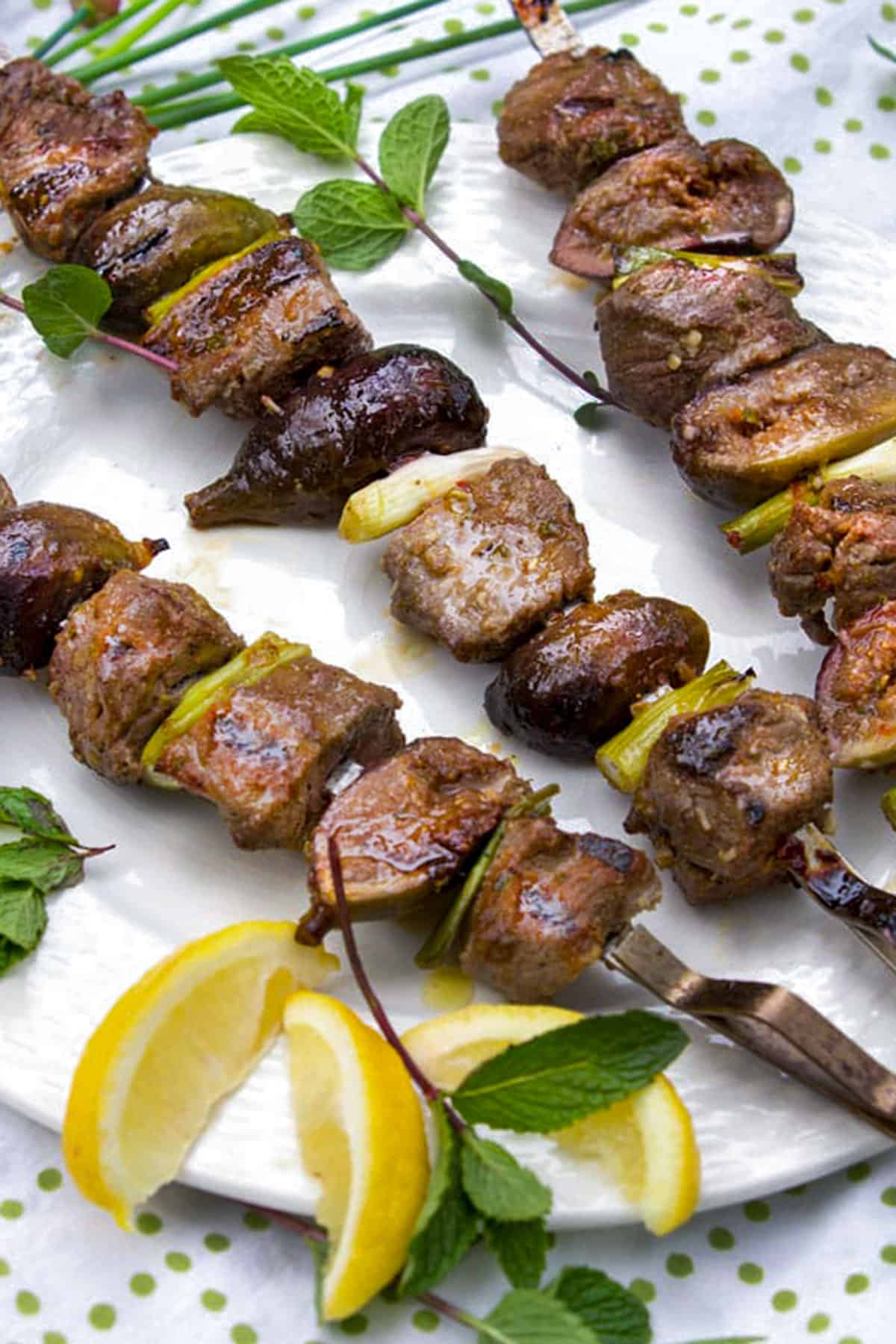 Three kebabs threaded with cubes of lamb, halved fresh figs and pieces of scallions, surrounded by lemon slices and mint sprigs