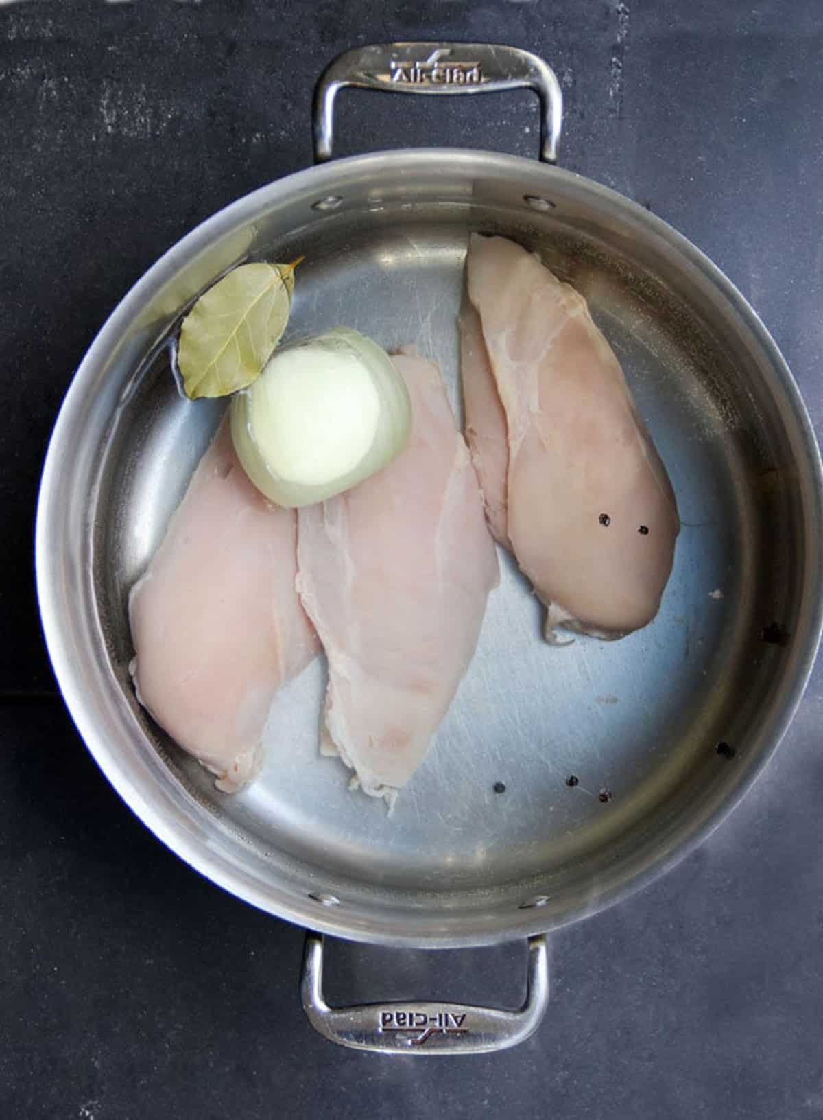 overhead shot of a large stainless steel saucepan filled with water and three raw chicken breasts and a half an onion and a bay leaf and some peppercorns and cloves