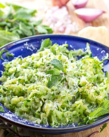 Blue bowl filled with sautéed shredded zucchini. A mint leaf on top.