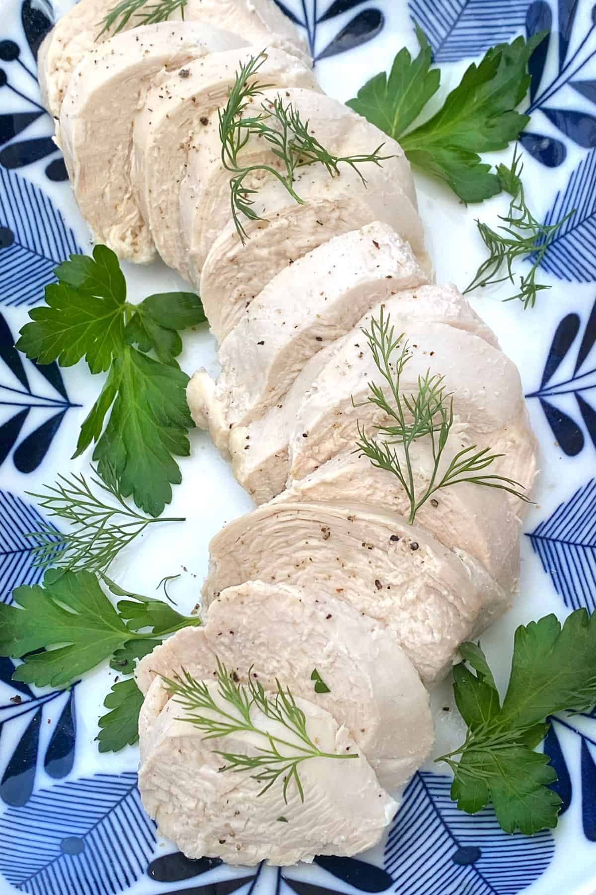 Sliced poached chicken on a blue and white platter with a few parsley and dill leave strewn around