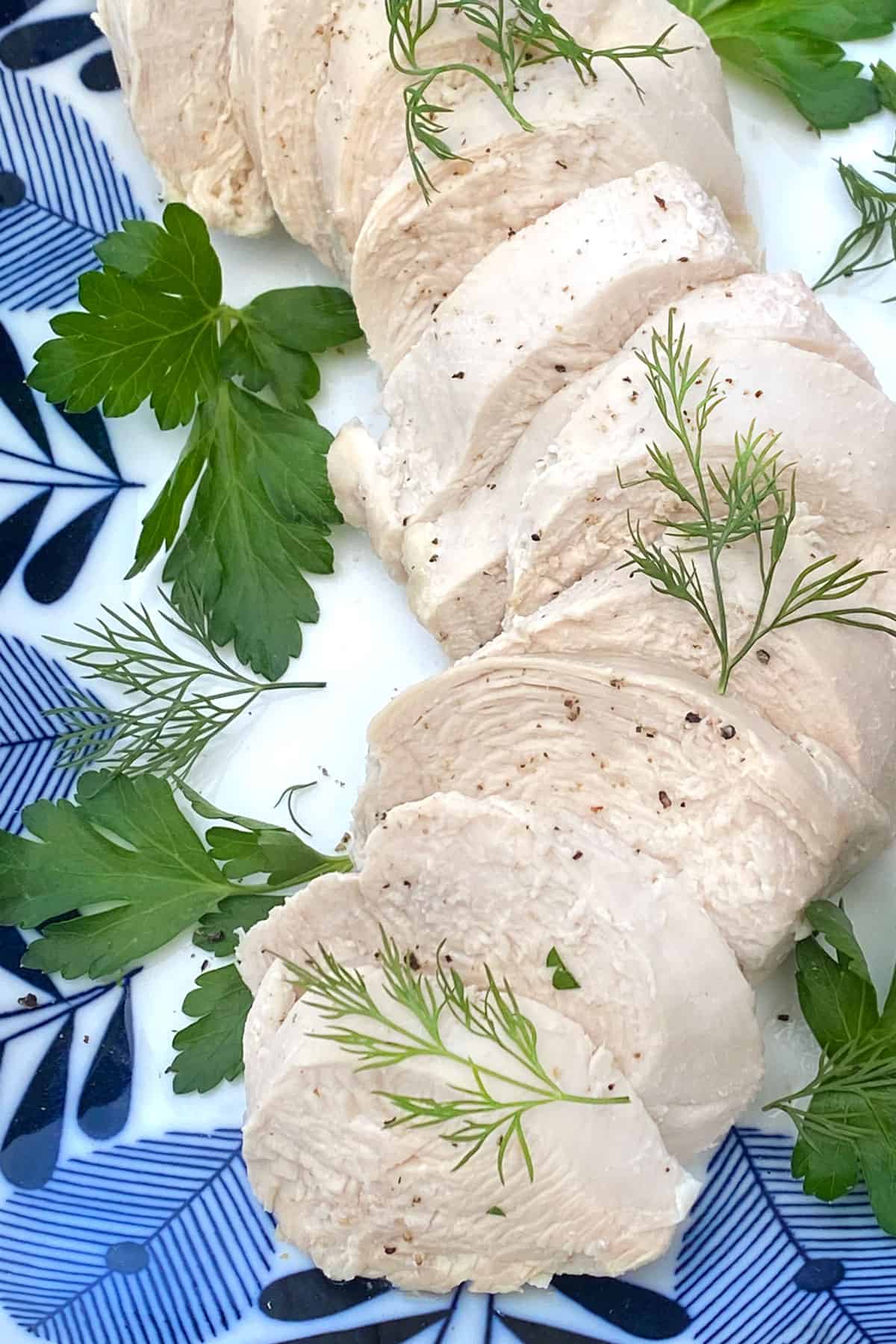 Close up of 5 slices of poached chicken on a blue and white plate, topped with some delicate sprigs of dill and parsley leaves