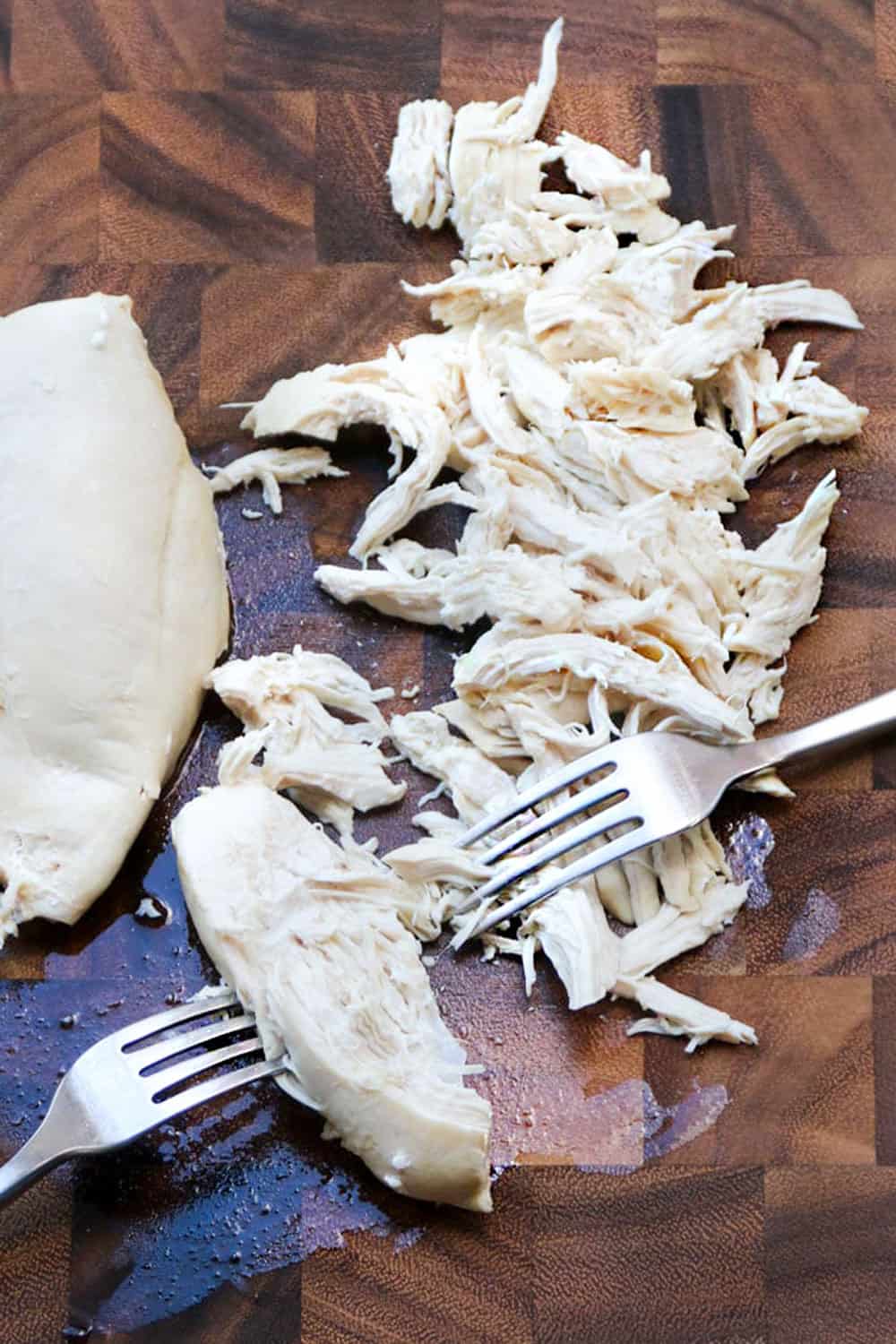 poached chicken being shredded using two forks, on a brown wooden cutting board, seen from above