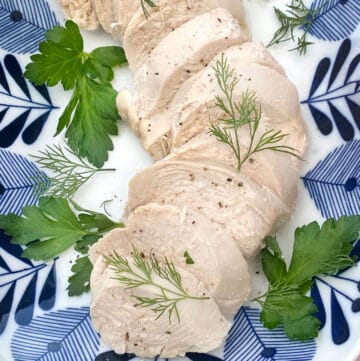 Sliced poached chicken on a blue and white platter with a few parsley and dill leave strewn around