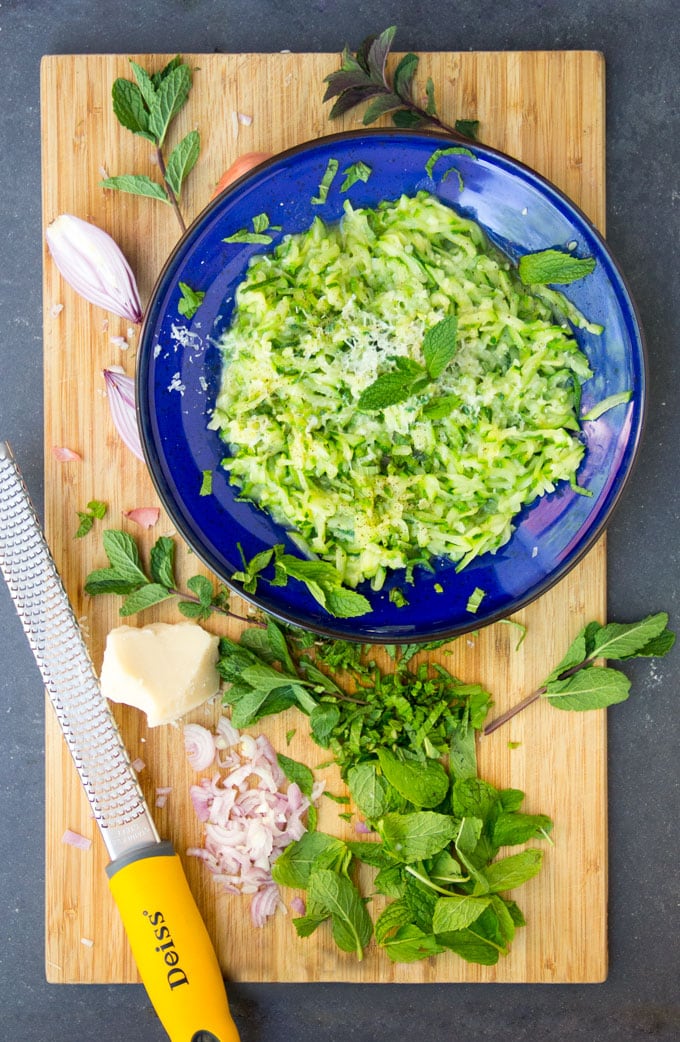 cutting board with mint leaves, a hunk of parmesan, a cheese grater and a blue bowl filled with sautéed shredded zucchini.