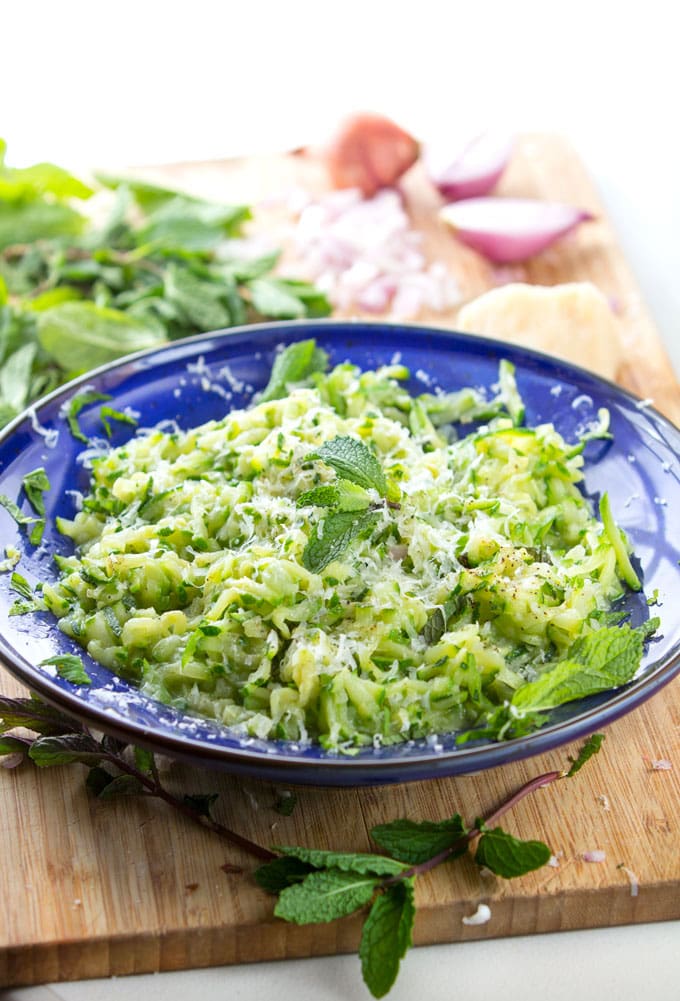 Blue bowl filled with shredded zucchini and a garnish of parmesan and basil, on a cutting board with chopped shallots and basil leaves