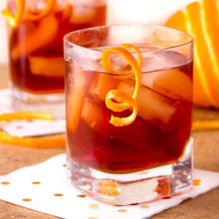 A vivid red cocktail in a low ball glass on ice with a swirly garnish of orange peel.