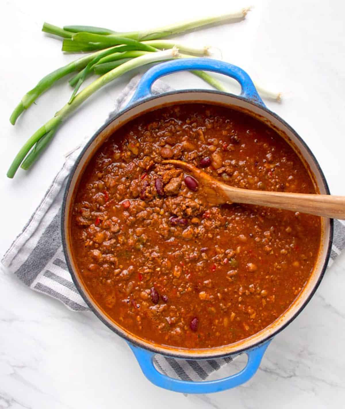 Dutch oven filled with chili con carne, a wooden spoon stuck into the chili and a few scallions on the counter beside the pot.