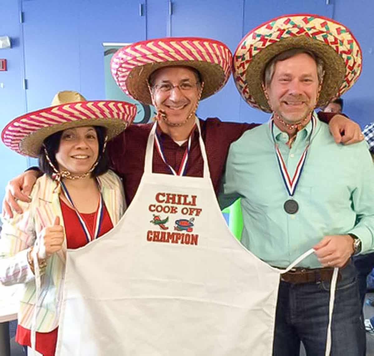 Ed Goldfinger, chili cook-off winner, wearing a sombrero and a chili winner apron, standing with second place winner Dean and another co-worker who took third place.