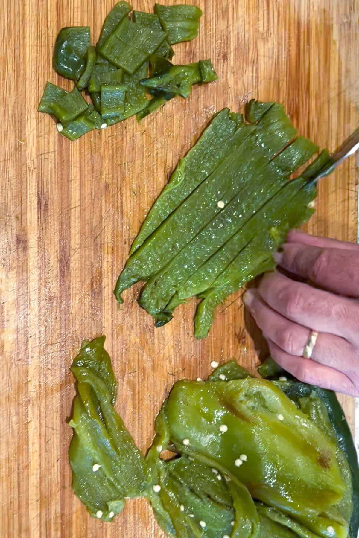 Poblano chilies on a wooden cutting board being sliced into strips, with some that have been diced on the side.