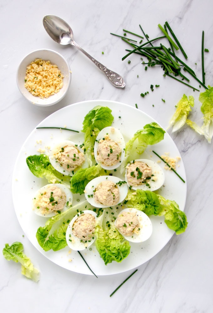 7 French Deviled Eggs on a white plate, decorated with baby romaine leaves, chopped egg yolks in a small bowl, chopped chives in the background