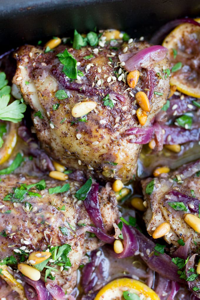 Close up of a few roast chicken thighs topped with colorful lemon slices, parsley, red onions, pine nuts and za'atar