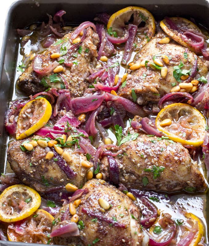 Baking pan with 5 cooked chicken thighs topped with lemon, red onions, pine nuts and spices.