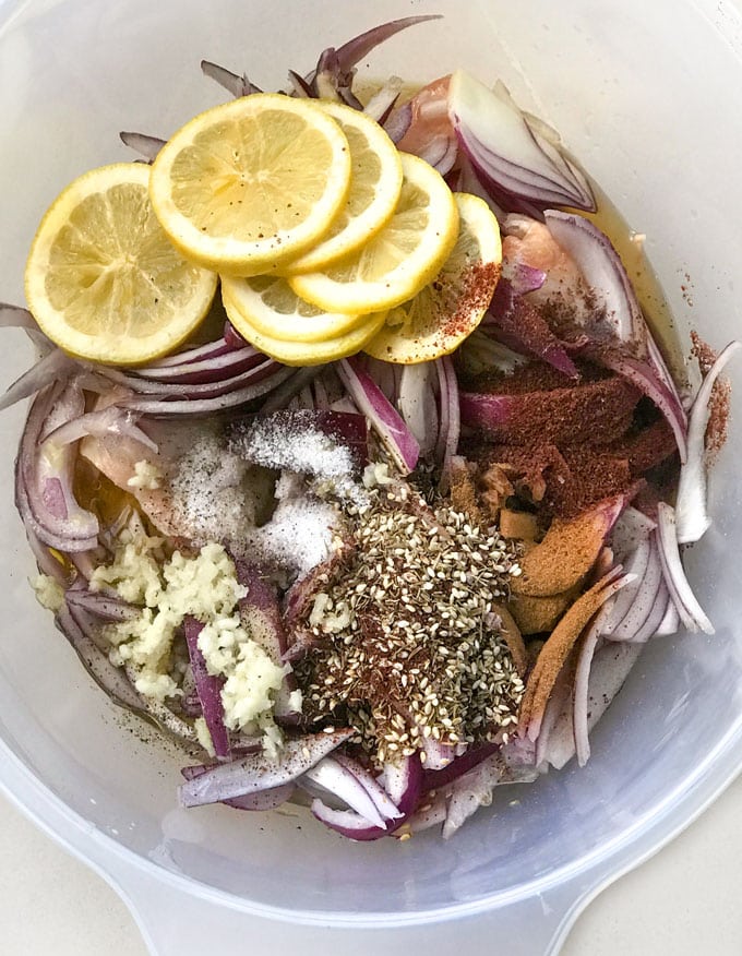plastic bowl filled with chicken thighs, red onions, spices, lemons and garlic.