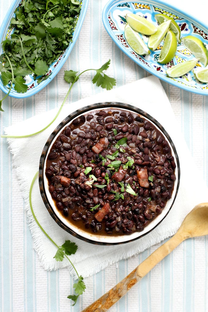 A bowl of Cuban black beans on a blue and white striped table cloth with a bowl of cilantro and bowl of lime wedges