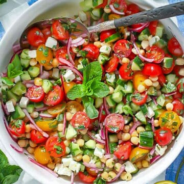 close up overhead shot of a white serving bowl filled with a colorful salad of sliced cherry tomatoes, chickpeas, cubed feta, chopped cucumbers, and mint leaves