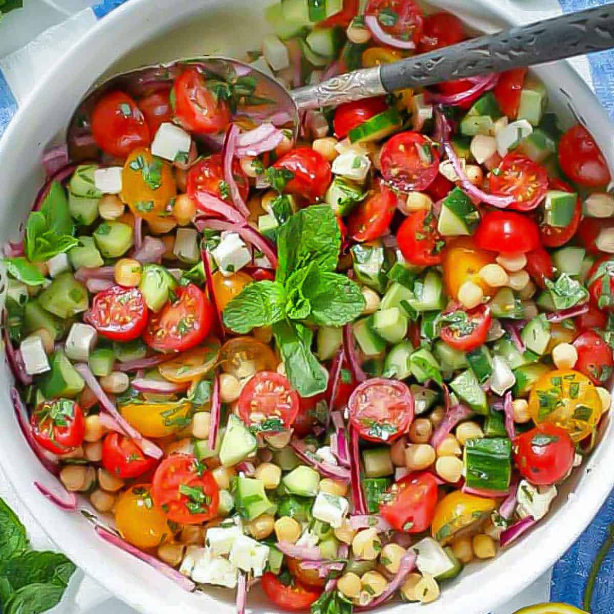 close up overhead shot of a white serving bowl filled with a colorful salad of sliced cherry tomatoes, chickpeas, cubed feta, chopped cucumbers, and mint leaves