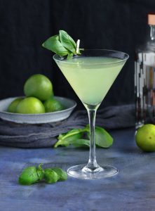 Basil vodka gimlet cocktail in a martini glass with two basil leave attached by a tiny clothespin, a bowl of limes in the background and a few basil leaves scattered on the blue talble