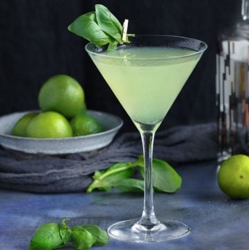 Basil vodka gimlet cocktail in a martini glass with two basil leave attached by a tiny clothespin, a bowl of limes in the background and a few basil leaves scattered on the blue talble