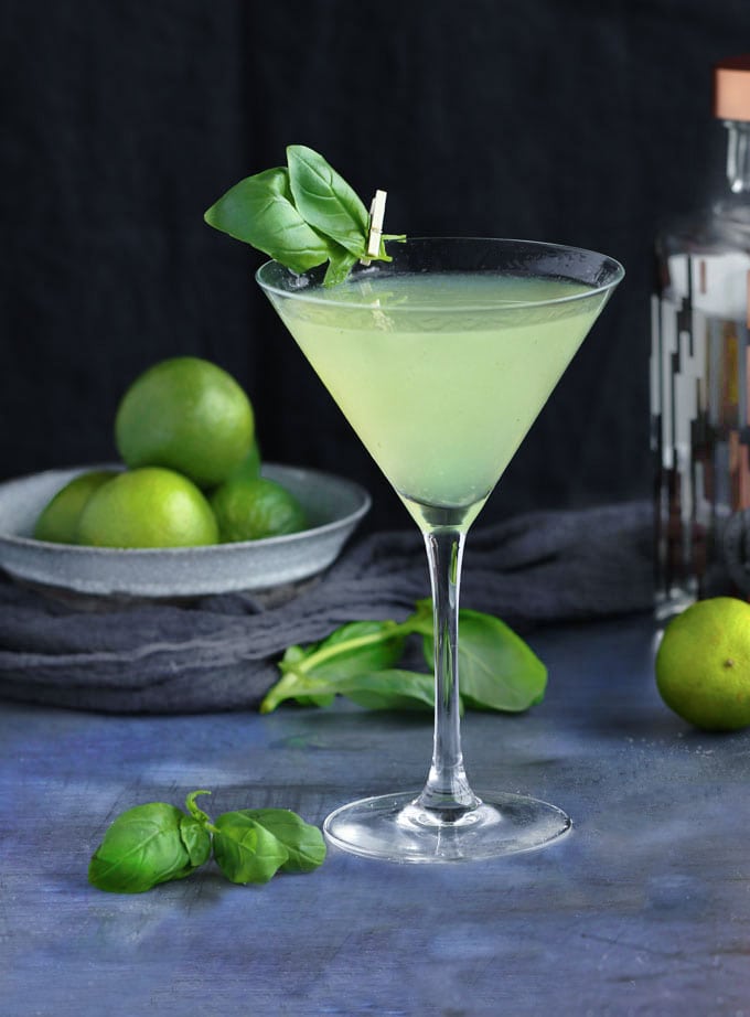 a martini glass filled with a green colored basil vodka gimlet and garnished with basil leaves attached to the glass with a mini clothespin. 