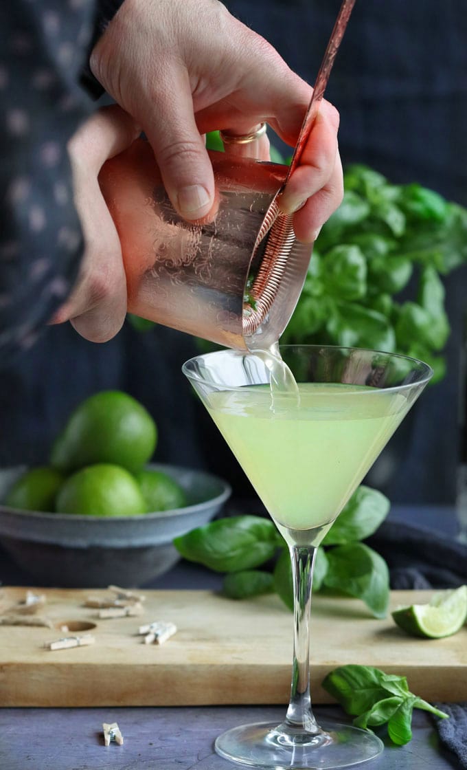 Hands pouring a basil vodka gimlet cocktail into a martini glass with lots of limes and basil leaves in the background