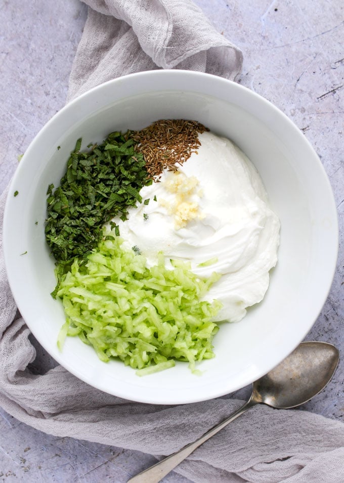 Little piles of ingredients in a white bowl: plain yogurt, grated cucumber, chopped mint leaves, toasted cumin seeds and grated garlic