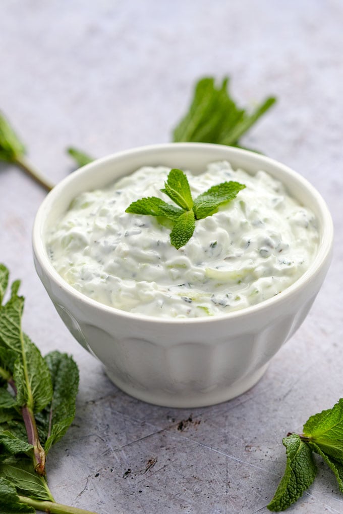 White bowl filled with white cucumber raita with a mint sprig in the center, on a lavender surface with mint sprigs strewn around