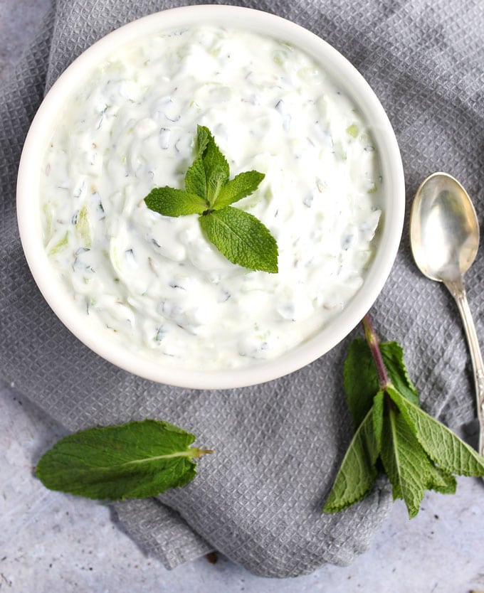 White bowl filled with white cucumber raita, flecked with mint and garnished with a sprig of fresh mint