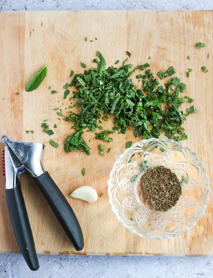 ingredients on a blond wood cutting board: chopped mint, cumin seeds in a small glass bowl, a clove of garlic and a garlic press