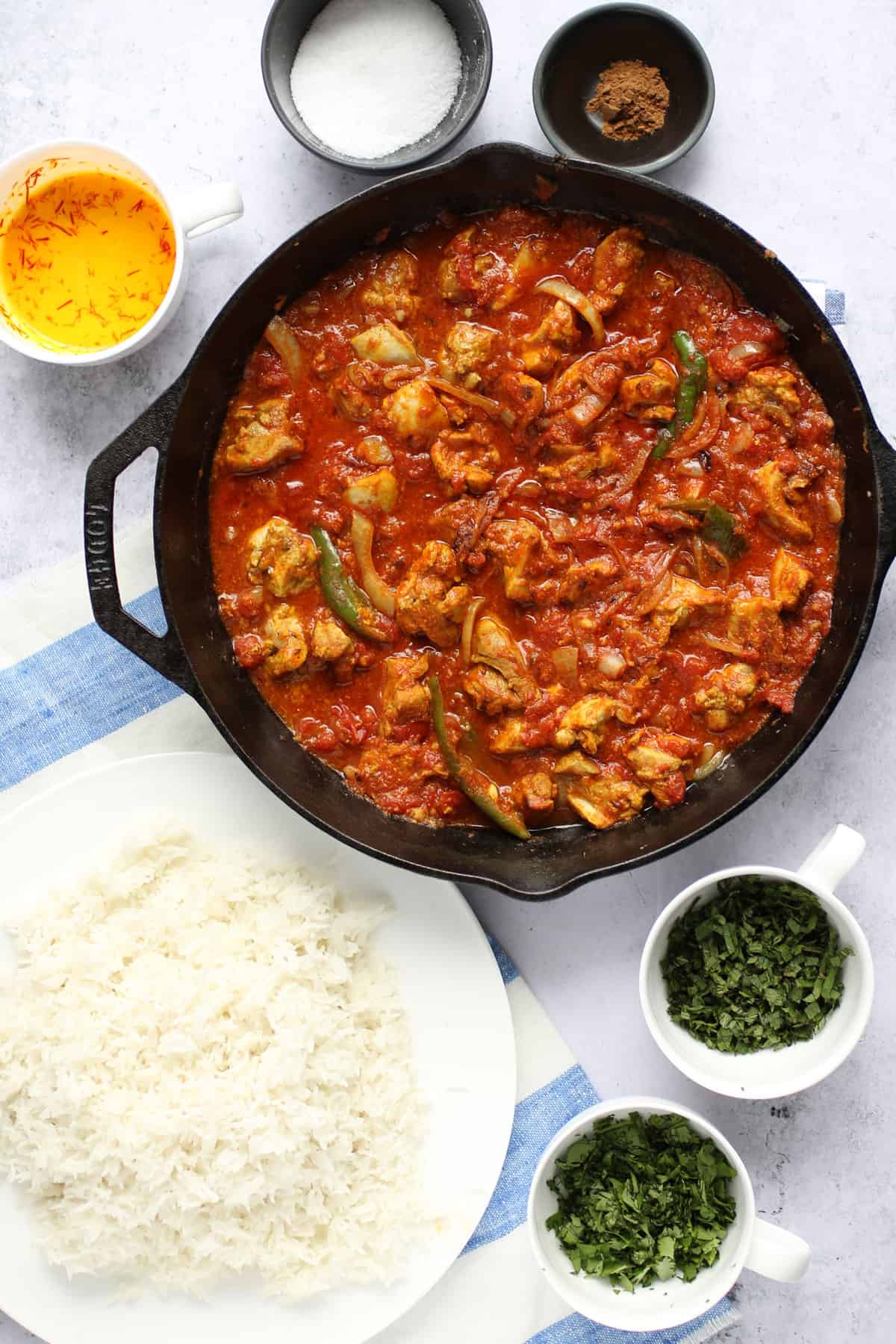 cast iron skillet filled with chicken curry surrounded by a large shallow bowl of cooked rice and small cups of chopped herbs, saffron milk, salt and garam masala