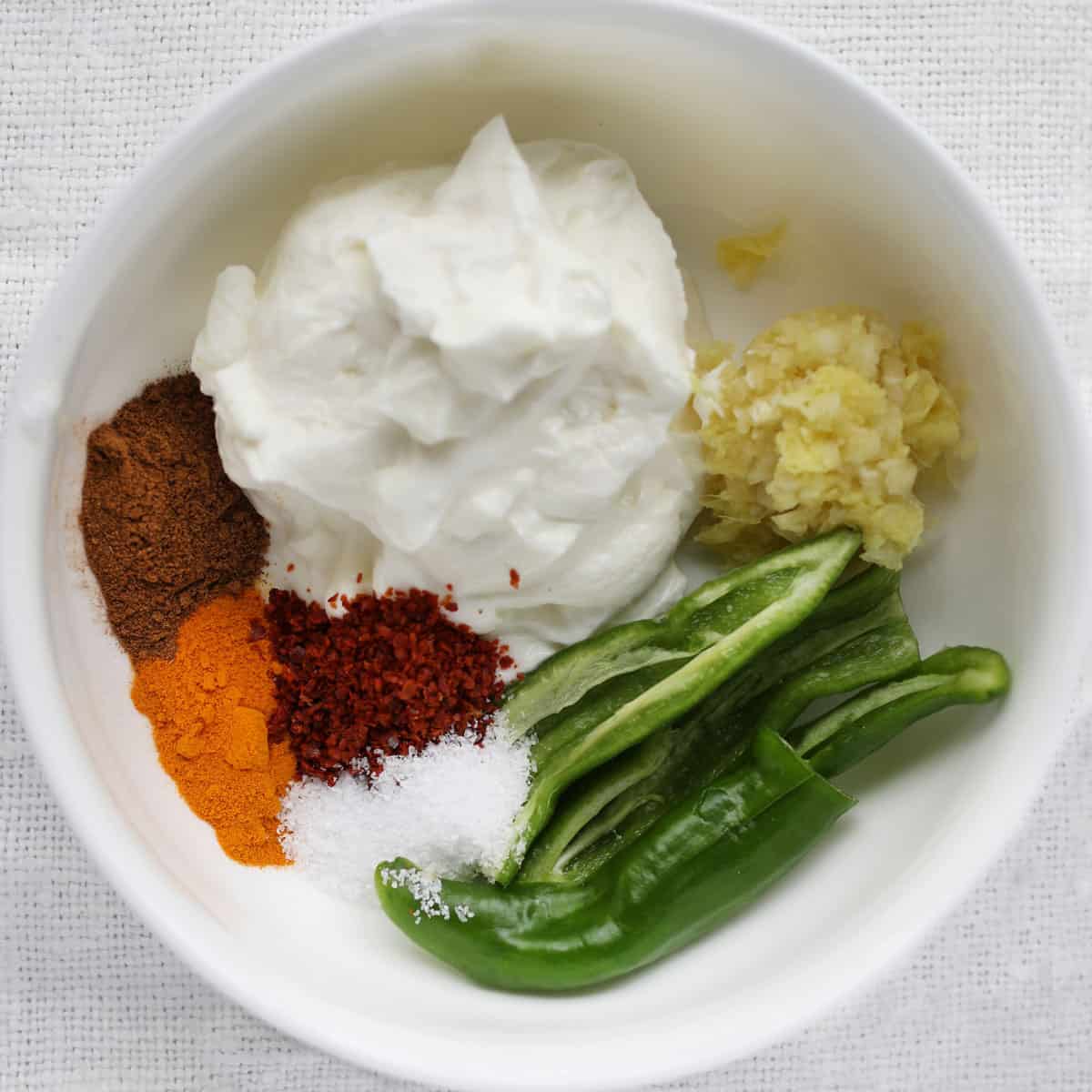 A plastic mixing bowl filled with separate marinade ingredients, yogurt, ginger-garlic paste, sliced green chilies, garam masala, chili powder and turmeric.