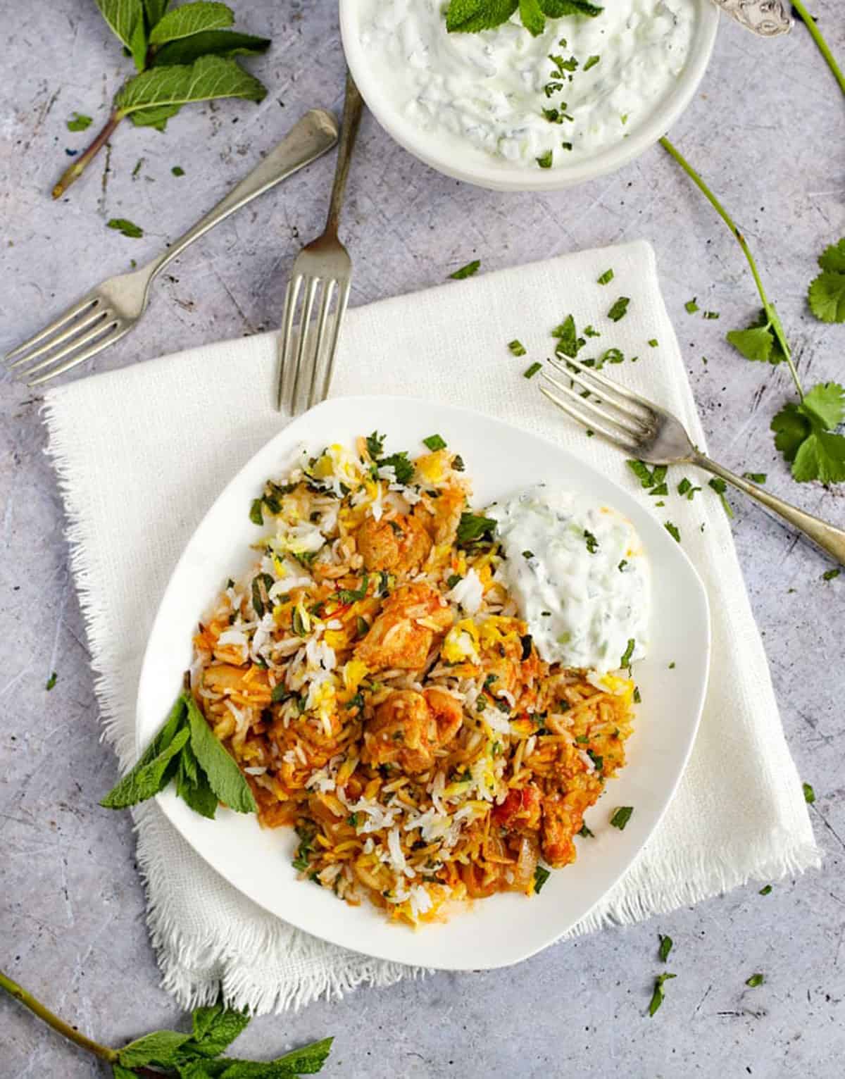 chicken biryani on a plate with forks on the table pointing to it and a bowl of raita next to it.