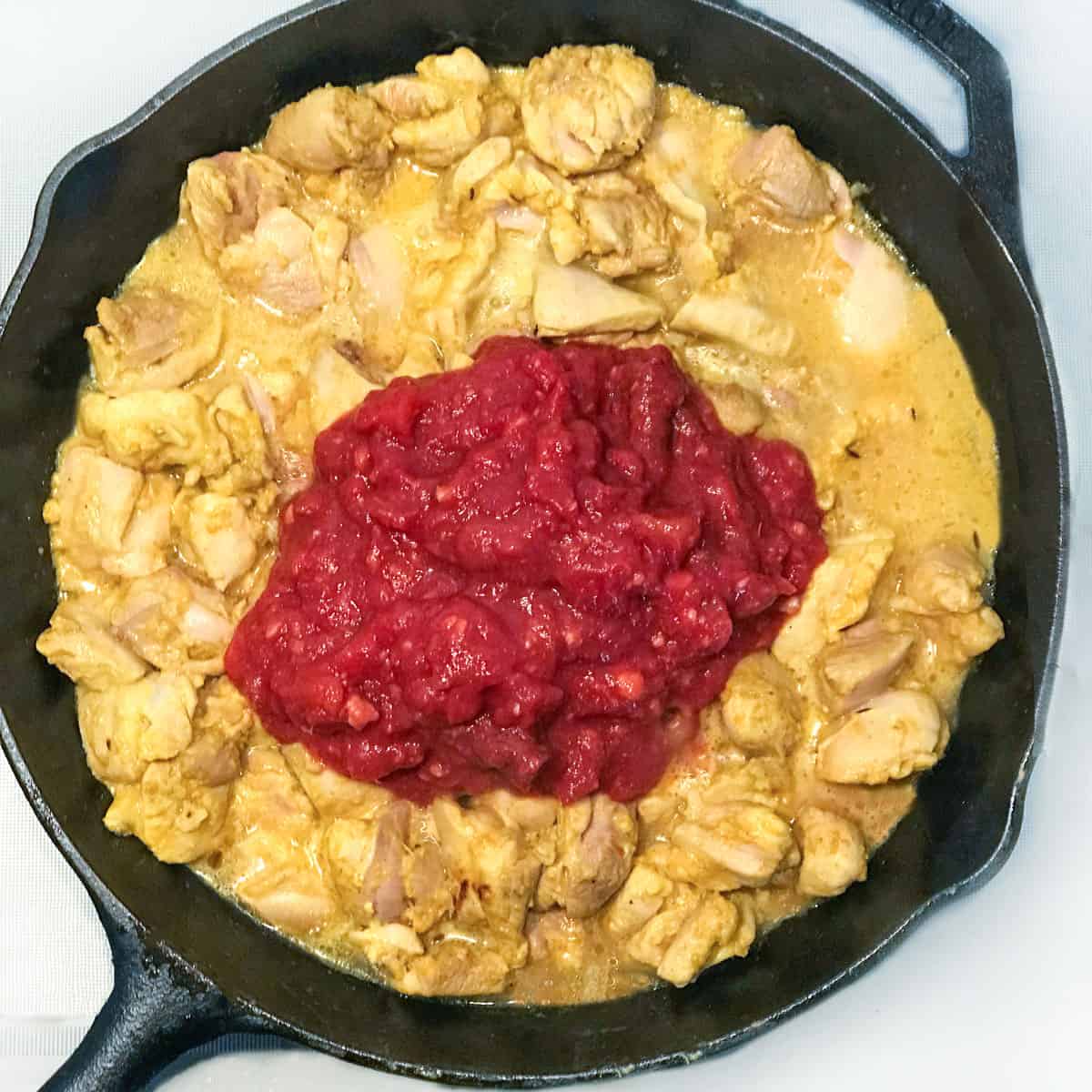 cubes of chicken and yogurt marinade simmering in a cast iron skillet and topped with chopped tomatoes