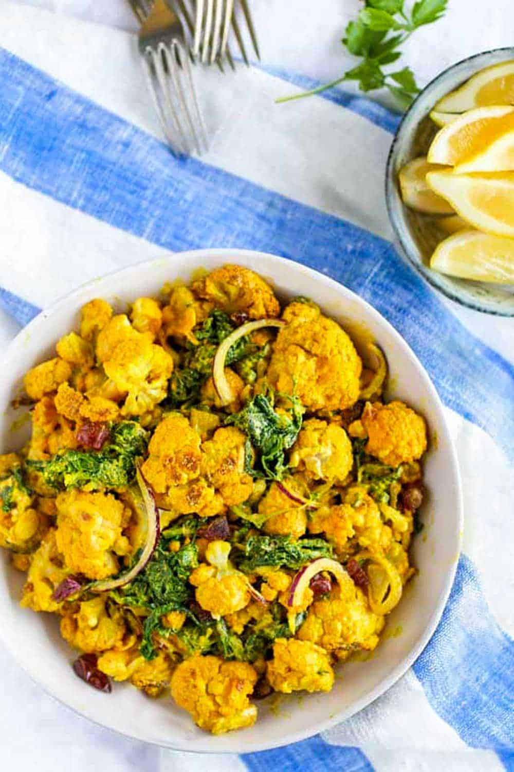 bright yellow turmeric cauliflower salad in a white bowl on a blue and white striped napkin with forks in the background