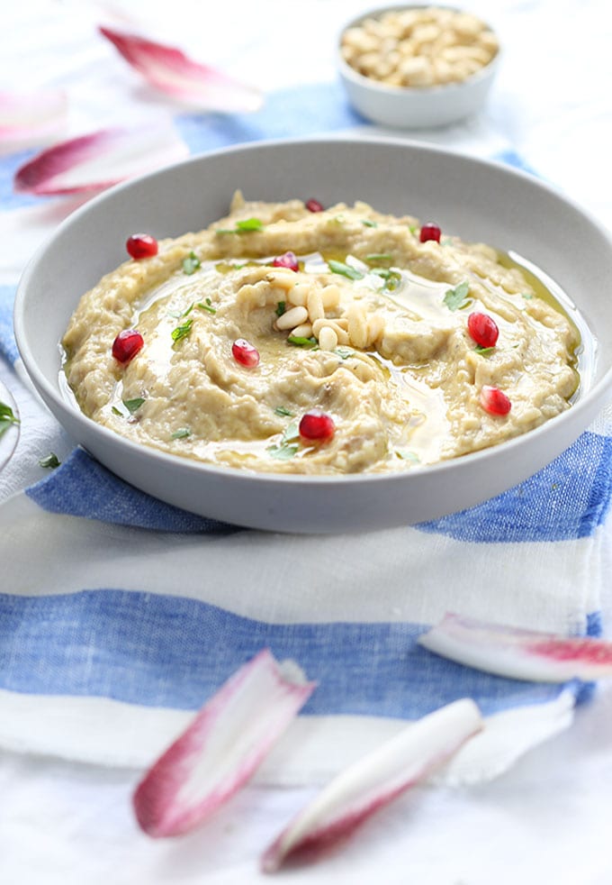 a bowl of baba ganoush garnished with pomegranate seeds, pine nuts, parsley and chicory leaves.