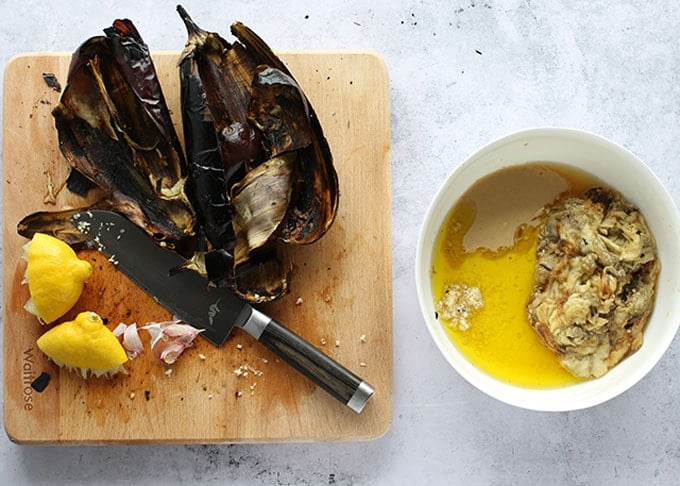 Ingredients and process shots for how to make baba ganoush at home: charred eggplant skins, lemon rinds, garlic skins on a cutting board and a bowl with eggplant flesh, olive oil, tahini, lemon juice and garlic. 