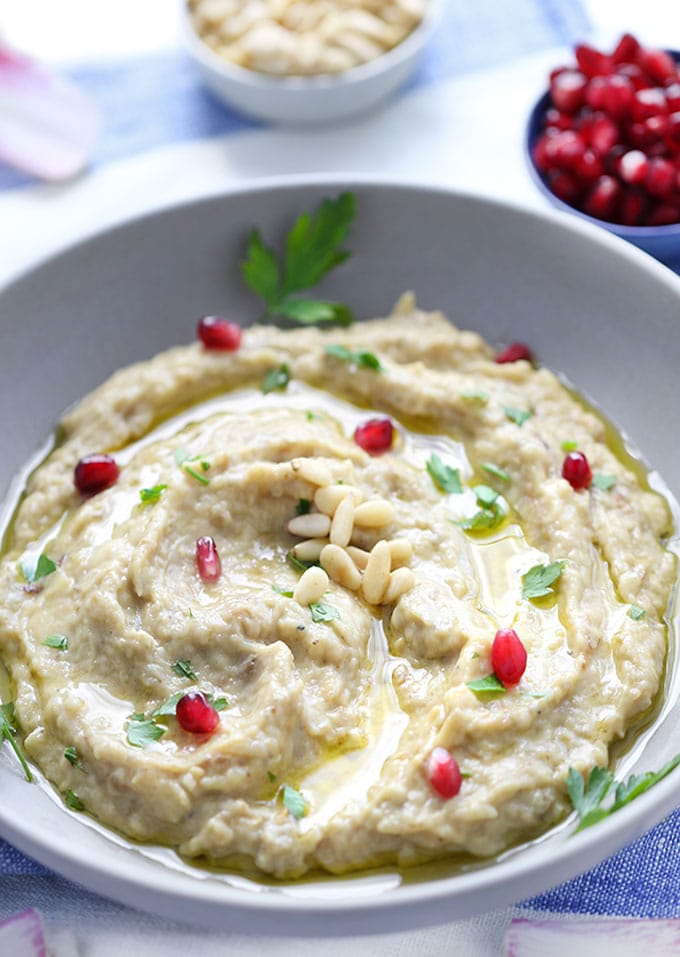 a bowl of baba ganoush garnished with pomegranate seeds, pine nuts, parsley and chicory leaves.