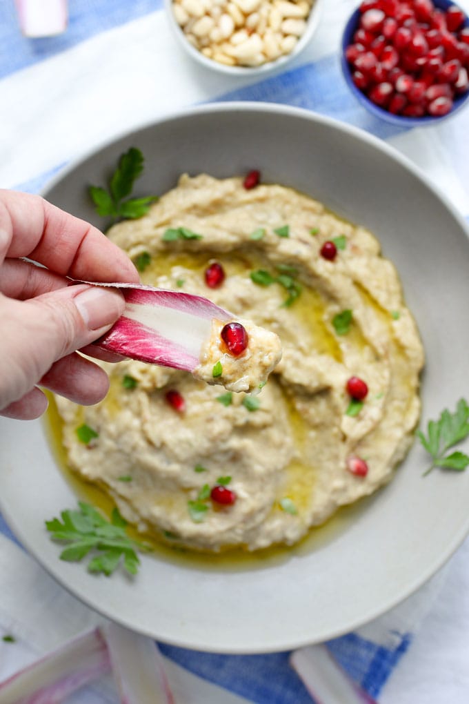A hand dipping a chicory leaf into a bowl of baba ganoush garnished with pomegranate seeds, pine nuts, parsley and chicory leaves.