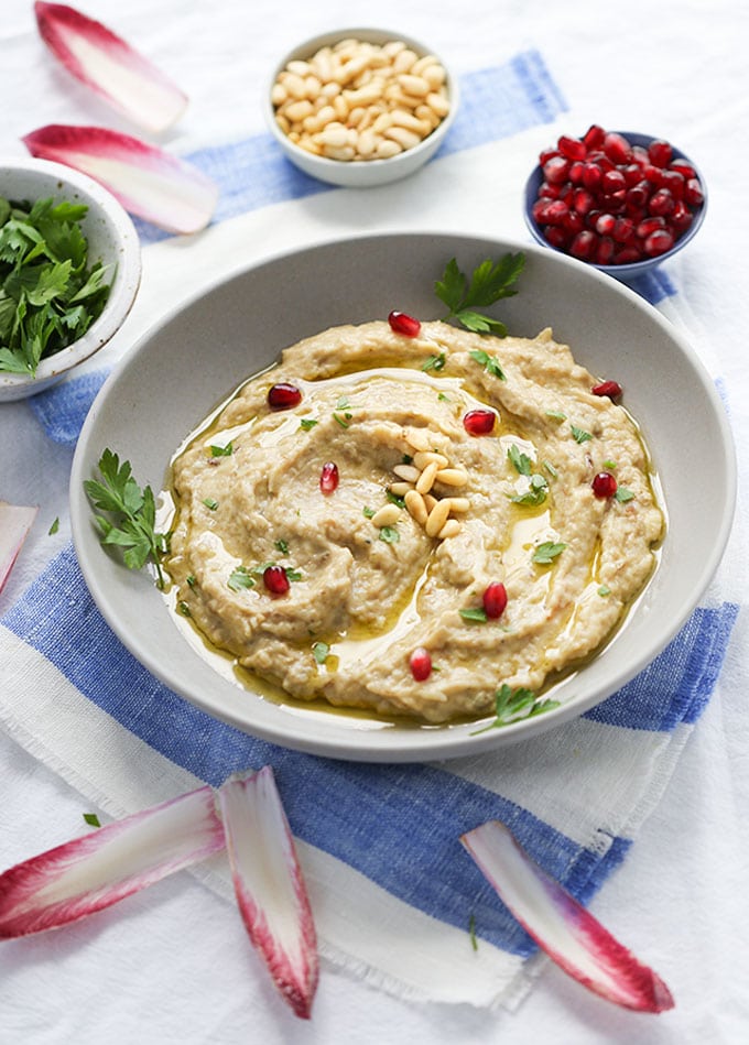 a bowl of baba ganoush garnished with pomegranate seeds, pine nuts, parsley and chicory leaves.