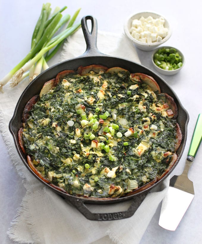 Greek spinach pie cooked in a skillet, topped with chopped scallions, accompanied by cubed feta.