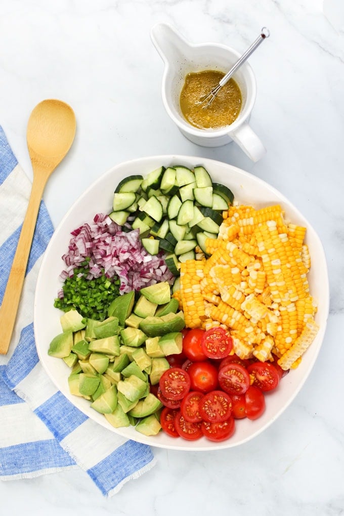 Ingredients for corn tomato avocado salad: Dressing plus a bowl with corn kernels, cubed avocado, sliced cucumbers, halved cherry tomatoes, chopped red onion and jalapeno peppers.