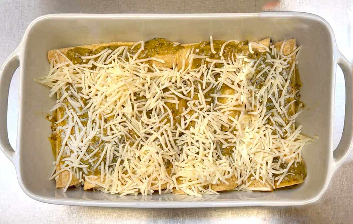 overhead shot of a white casserole dish filled with a dozen rolled chicken enchiladas topped with salsa verde and shredded cheese