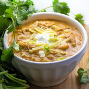 Bowl of creamy white bean and chicken chili topped with a blog of sour cream in the middle and shredded white cheddar. Sprigs of cilantro in the background.