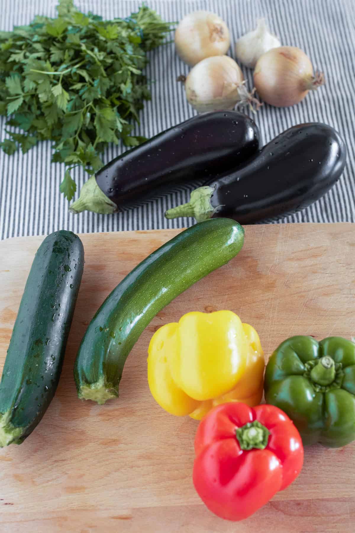 two eggplants, two zucchinis, three bell peppers, a few yellow onions, a bulb of garlic and a bunch of parsley - on a kitchen counter with a wooden cutting board