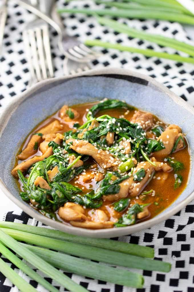 a blue bowl on a black and white table cloth, filled with a stew of chicken and spinach in Korean gochujang sauce.