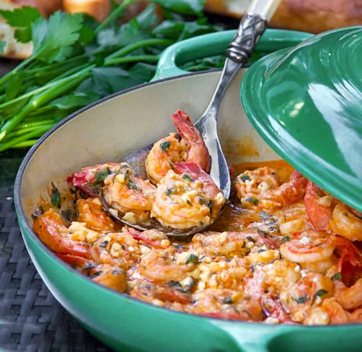 Shallow dutch oven with the lid cracked open showing a pot of lemon butter garlic shrimp with feta and a spoon scooping out a few shrimp