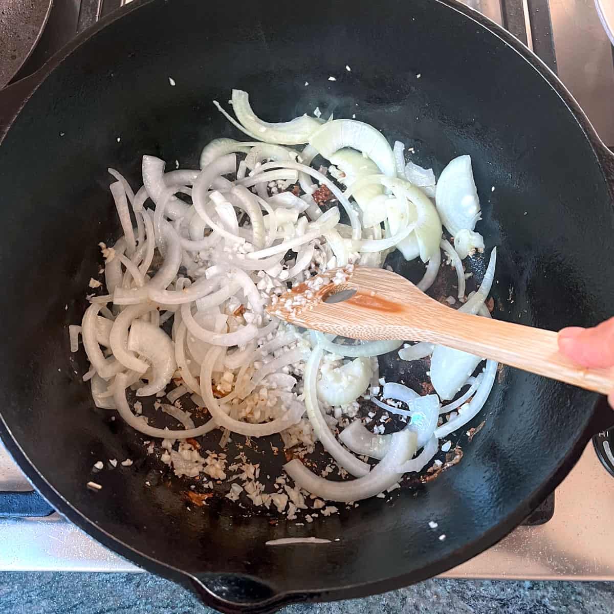 Thinly sliced onions and garlic cooking in a cast iron skillet, being stirred with a wooden spoon.