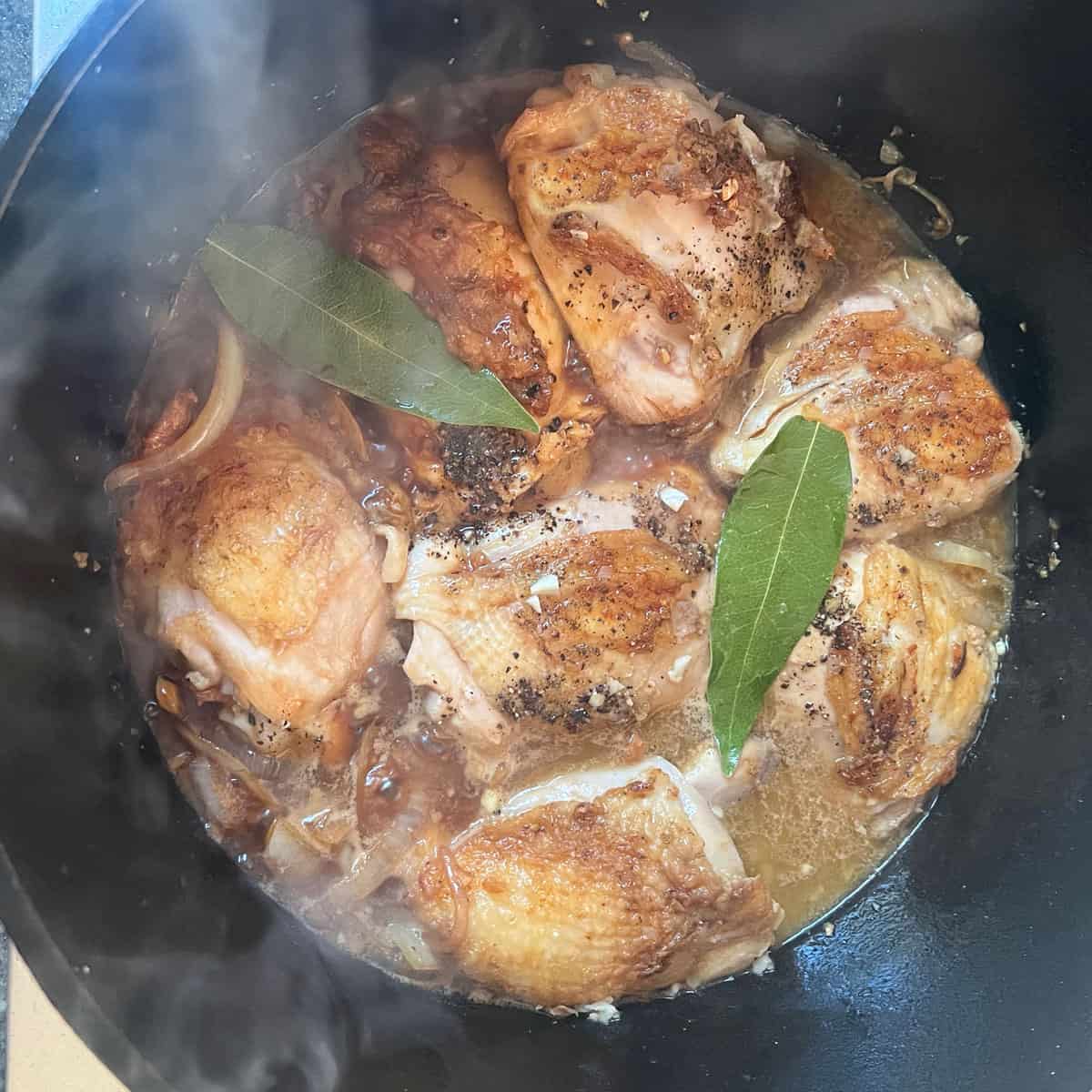 7 browned chicken thighs in a Dutch oven, surrounded with cooking liquid and topped with two bay leaves