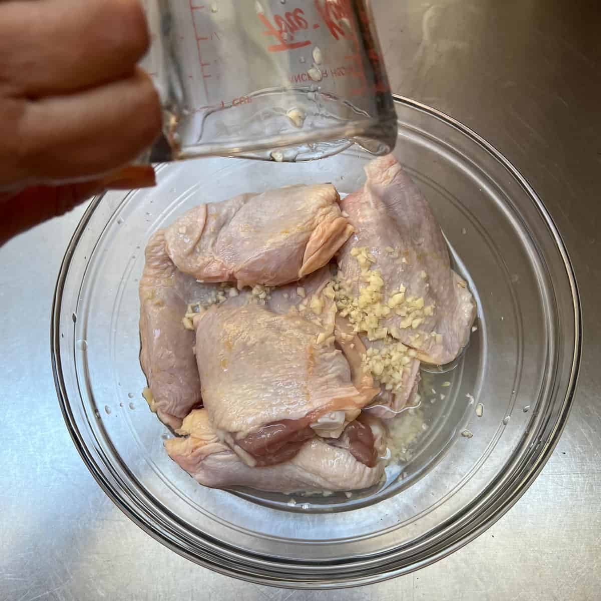 glass bowl filled with 8 raw chicken thighs, a measuring cup pouring white vinegar and water over them.