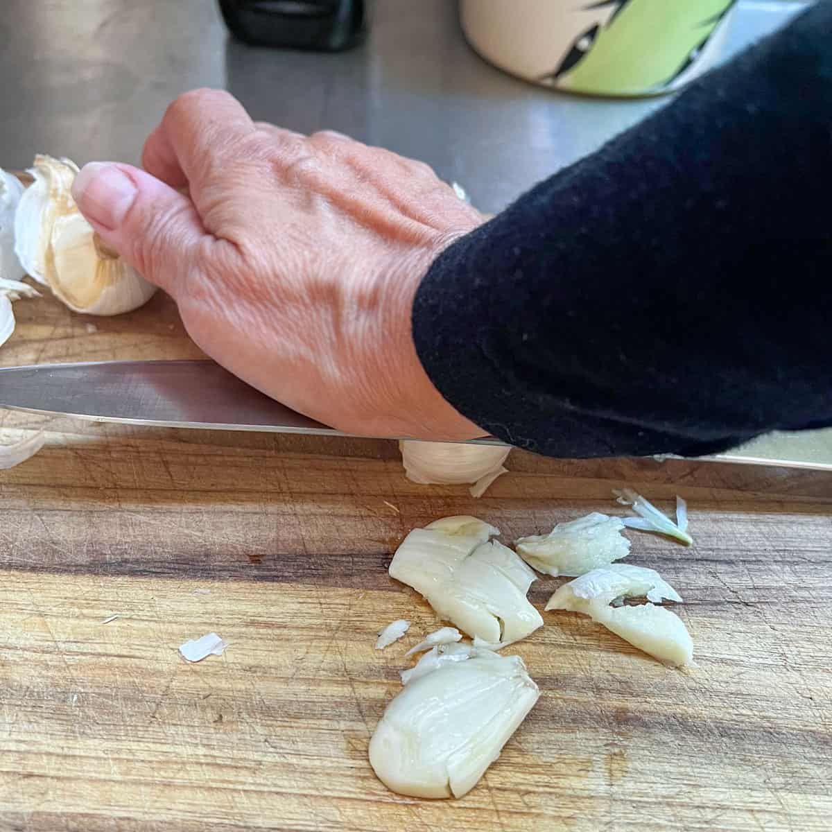 Someone's hand pressing down on the flat side of a chef's knife with a garlic clove underneath.